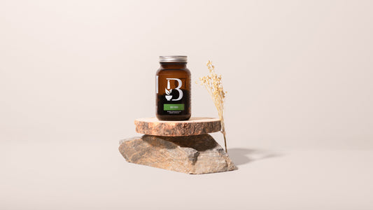 An amber glass supplement bottle with a silver cap and a black-and-white label featuring a large 'B' and green accent, displayed on a stacked wood slice and stone pedestal with dried grass against a warm beige background.