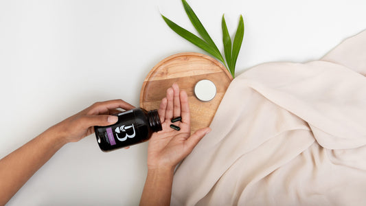 A person's hands are shown holding a bottle of Botanica Milk Thistle liquid capsules and pouring capsules into their palm, set against a clean white background with a beige blanket, a wooden tray, and green leaves.