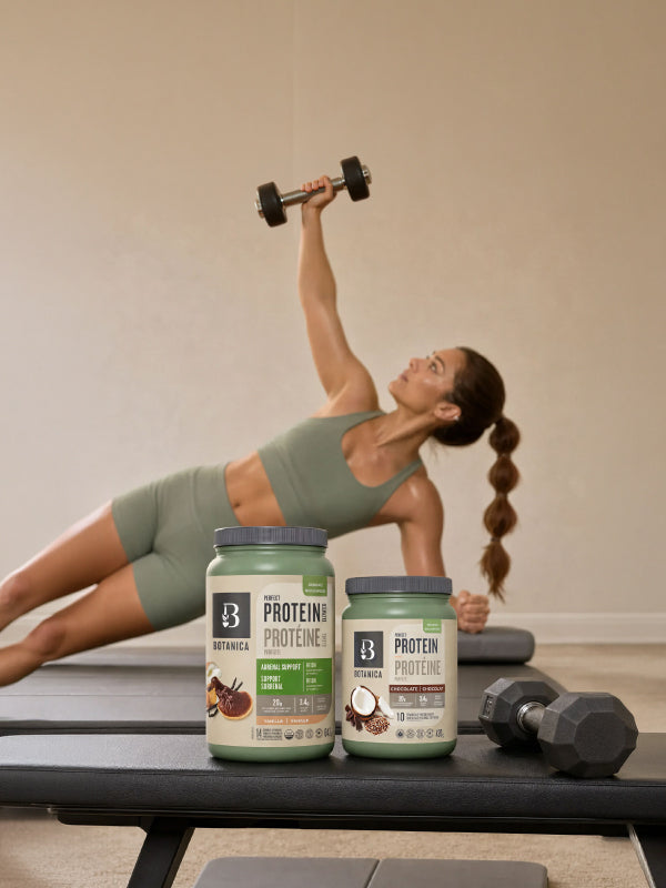 A woman in a green sports bra and shorts does a side plank exercise while holding a dumbbell overhead. In the foreground, two tubs of Botanica Perfect Protein Powder sit on a bench, with another dumbbell nearby, against a plain beige wall.