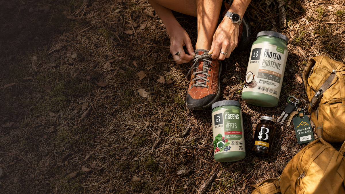 A top-down outdoor shot of a person tying the laces of an orange hiking boot while. Botanica products are laid out on the forest floor next to a tan backpack.