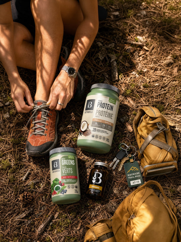 A top-down outdoor shot of a person tying the laces of an orange hiking boot while. Botanica products are laid out on the forest floor next to a tan backpack.
