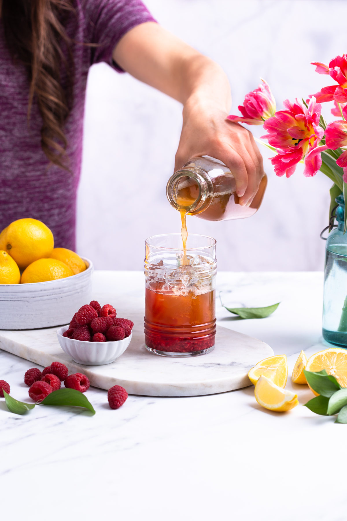 A woman's hand pouring Raspberry lemon-mint iced tea made with Botanica sweetened lion's mane iced tea with a bowl of lemons and a small ramekin of fresh raspberries and on a marble countertop