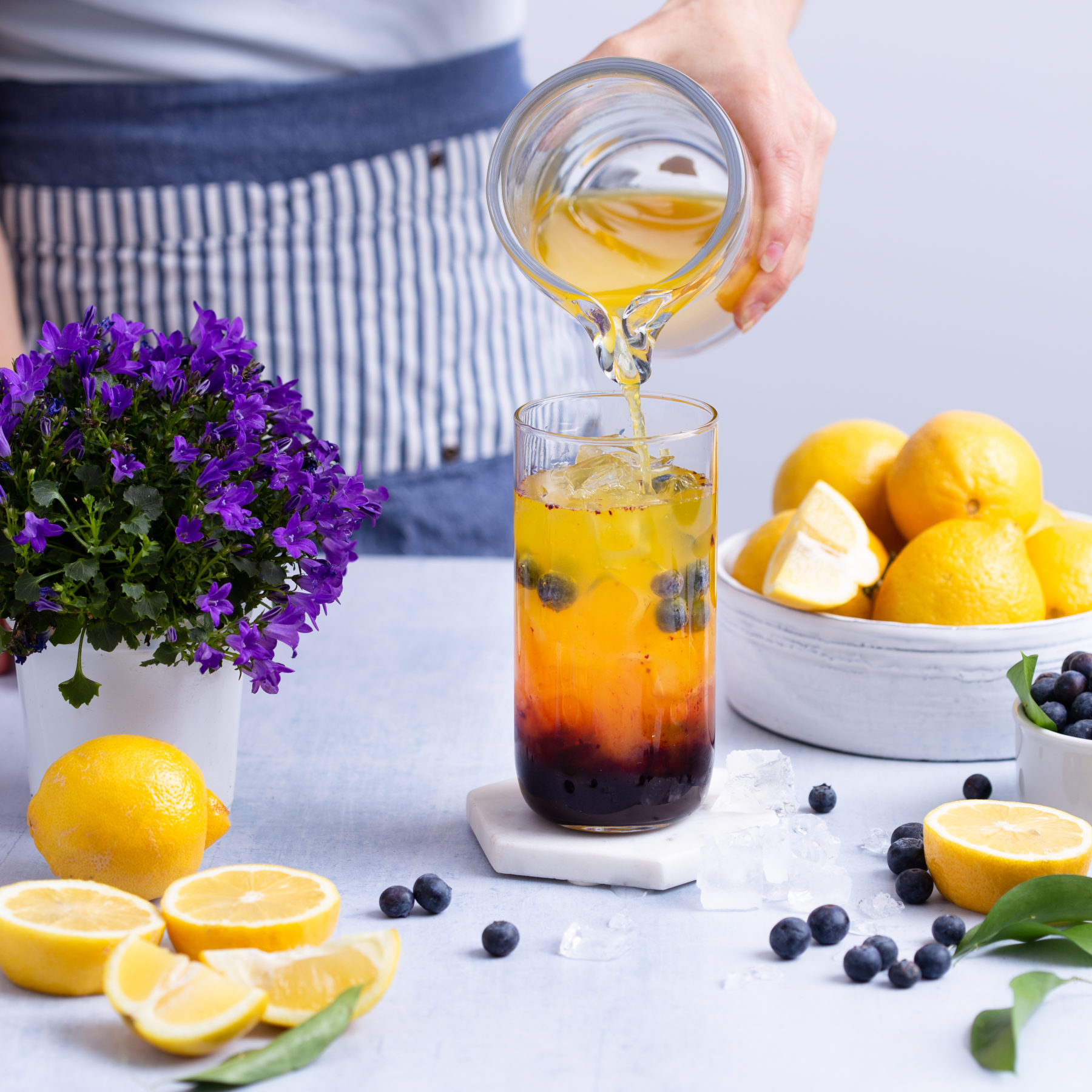Pouring a glass of blueberry lavender turmeric lemonade over ice from a pitcher onto a white marble table with purple flowers, a bowl of fresh lemons and blueberries scattered.