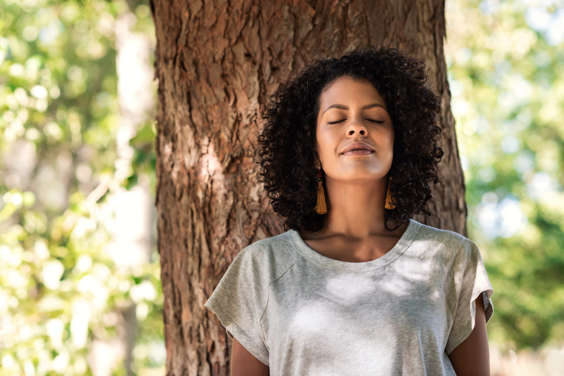 Peaceful young woman with curly hair leaning with her eyes closed against a tree trunk on a summer day